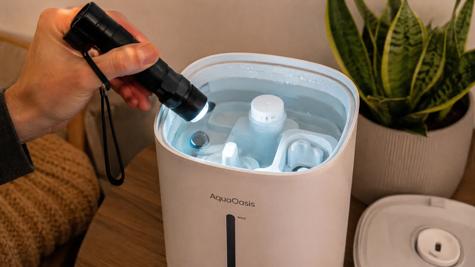 A hand holding a small flashlight up to the open tank of a white cool mist humidifier to check the inside for buildup