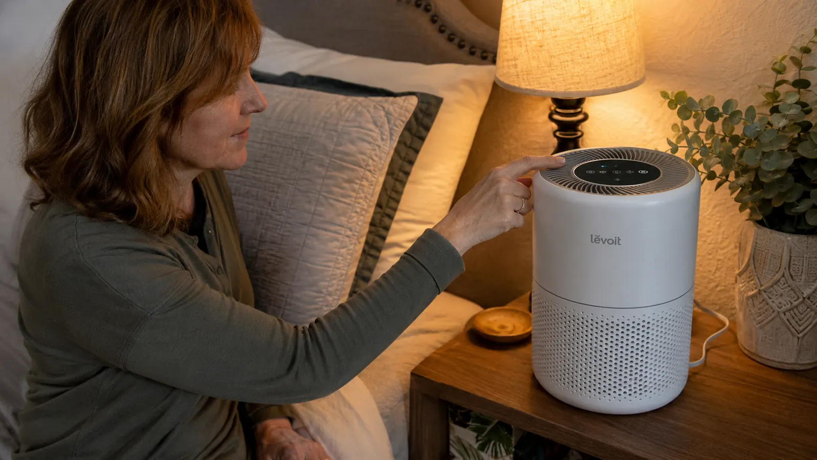 A hand pressing the sleep mode button on a white air purifier on a bedroom nightstand