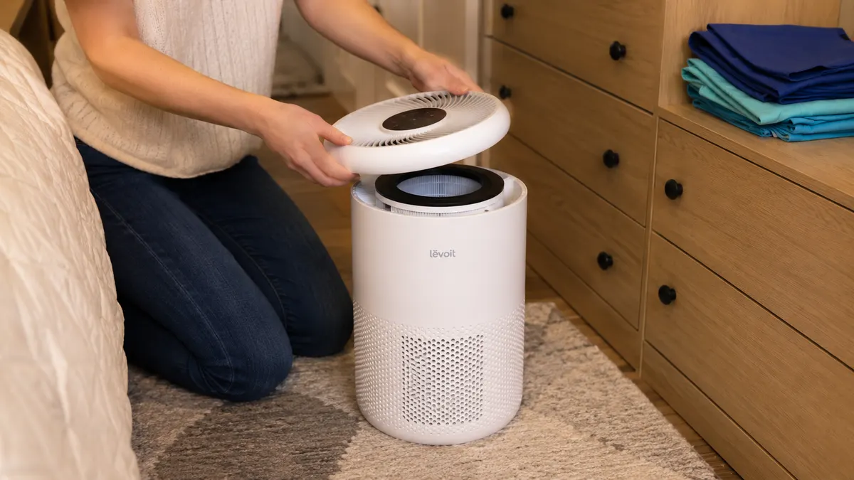Close-up of a hand lifting the top panel off a small white air purifier on a bedroom dresser to check the filter